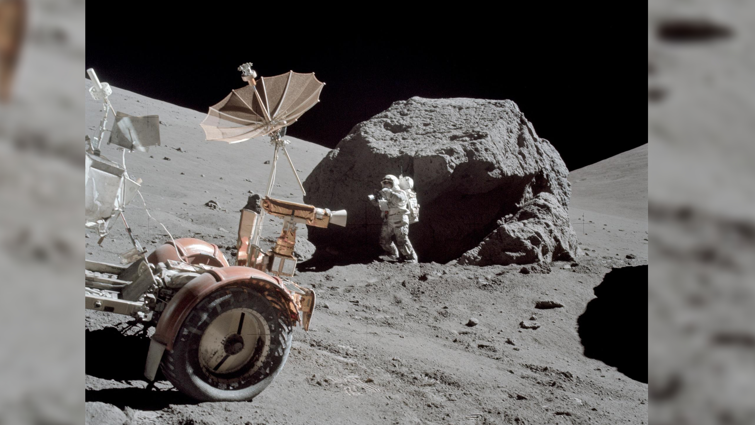 an astronaut stands on the surface of the moon. there is a large boulder in the background.