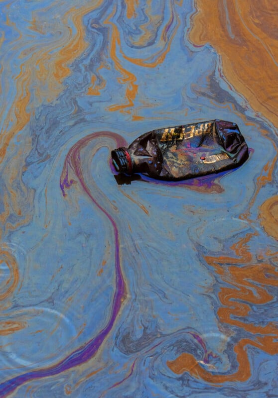 A crumpled plastic bottle floats on water with iridescent swirls of blue, brown, and orange, suggesting pollution or an oil spill.