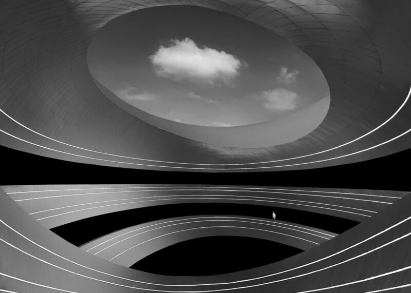 A person stands alone on a curved, futuristic structure with layered, spiral ramps and an open oval ceiling, revealing a blue sky with a single white cloud. The scene is in black and white.