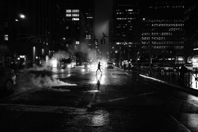 A person walks alone across a wet city street at night, illuminated by streetlights and surrounded by tall buildings. Steam rises from a vent, creating a moody, atmospheric scene in black and white.