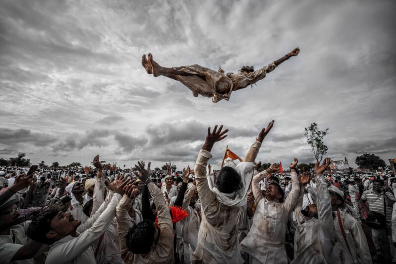A group of people dressed in white joyfully toss a person high into the air during a lively outdoor celebration, with dramatic clouds filling the sky above the large gathered crowd.