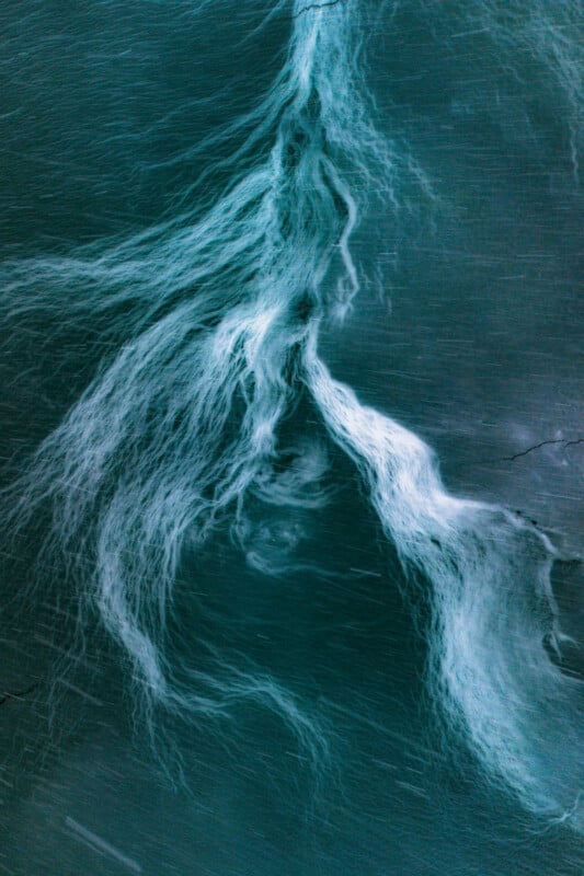 Aerial view of swirling white foam and streaks in deep blue-green water, creating an abstract, flowing pattern resembling marble or smoke.