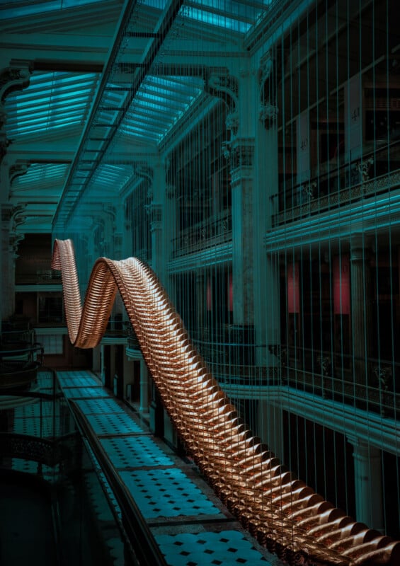 A large, illuminated, wave-shaped sculpture hangs in the atrium of a grand building with tall columns, glass ceilings, and patterned floors under teal lighting.