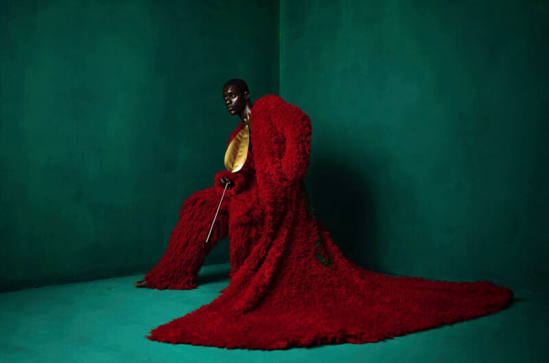 A person poses in a dramatic, long red textured coat against a green background. They sit on a stool, holding a cane and wearing a golden top, creating a striking contrast with the bold colors and luxurious fabrics.