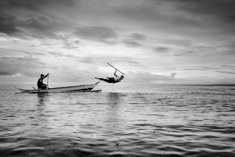 A black and white photo of a person paddling a small boat on the water, while another person leaps mid-air into the sea holding a stick, with clouds and horizon in the background.