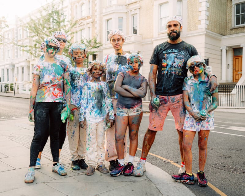 A group of seven people, including adults and children, stand on a city street covered in colorful paint splatters, looking at the camera. They wear goggles, shower caps, and casual clothes. Houses line the street behind them.