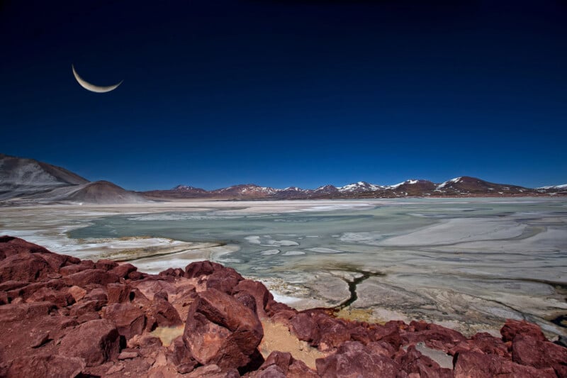 A crescent moon hangs in a deep blue sky over a barren, rocky desert landscape with red stones in the foreground and snow-capped mountains in the distance. Patches of salt flats and still water cover the ground.