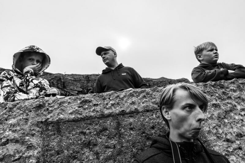 Four people stand outdoors behind a rough stone wall, all with serious or contemplative expressions. The sky is overcast, and the photo is black and white, emphasizing the mood and textures.