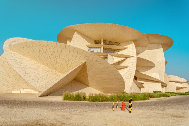 A modern beige building with large, overlapping, disk-shaped layers, resembling desert rose crystals, under a clear blue sky. Two yellow-black posts and a red hydrant are in the foreground.