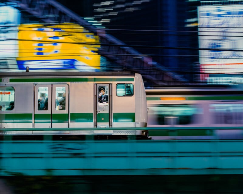 A train speeds by in an urban setting at night, with blurred neon signs in the background. Passengers can be seen inside, and a conductor stands in the doorway, illuminated by the train’s interior lights.