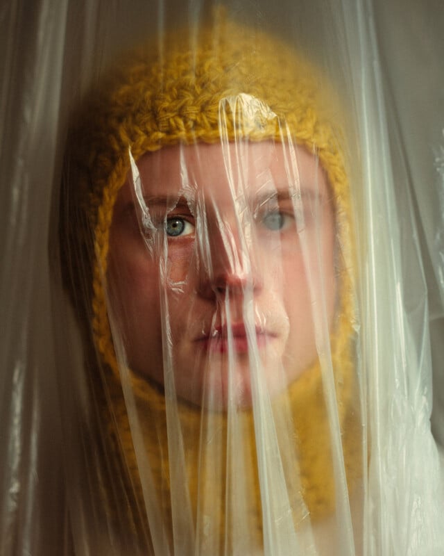 A person wearing a yellow knitted hood is seen through a sheet of translucent plastic, with their face partially blurred and distorted by the plastic.