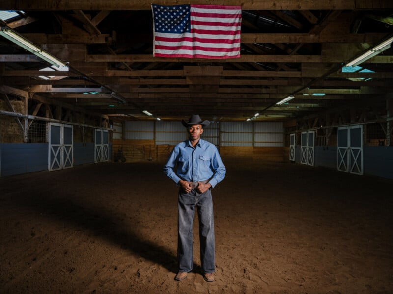 A man wearing a cowboy hat, denim shirt, and jeans stands confidently in the center of a barn with dirt floors. An American flag hangs above him on the wooden rafters. Light shines down, highlighting him.