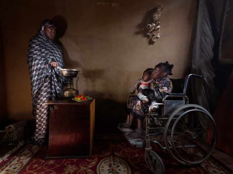 A woman in a patterned cloak stands by a scale on a small table. Beside her, a girl sits in a wheelchair holding a baby. Garlic hangs on the wall; vegetables are on the table, all in a dimly lit, modest room.