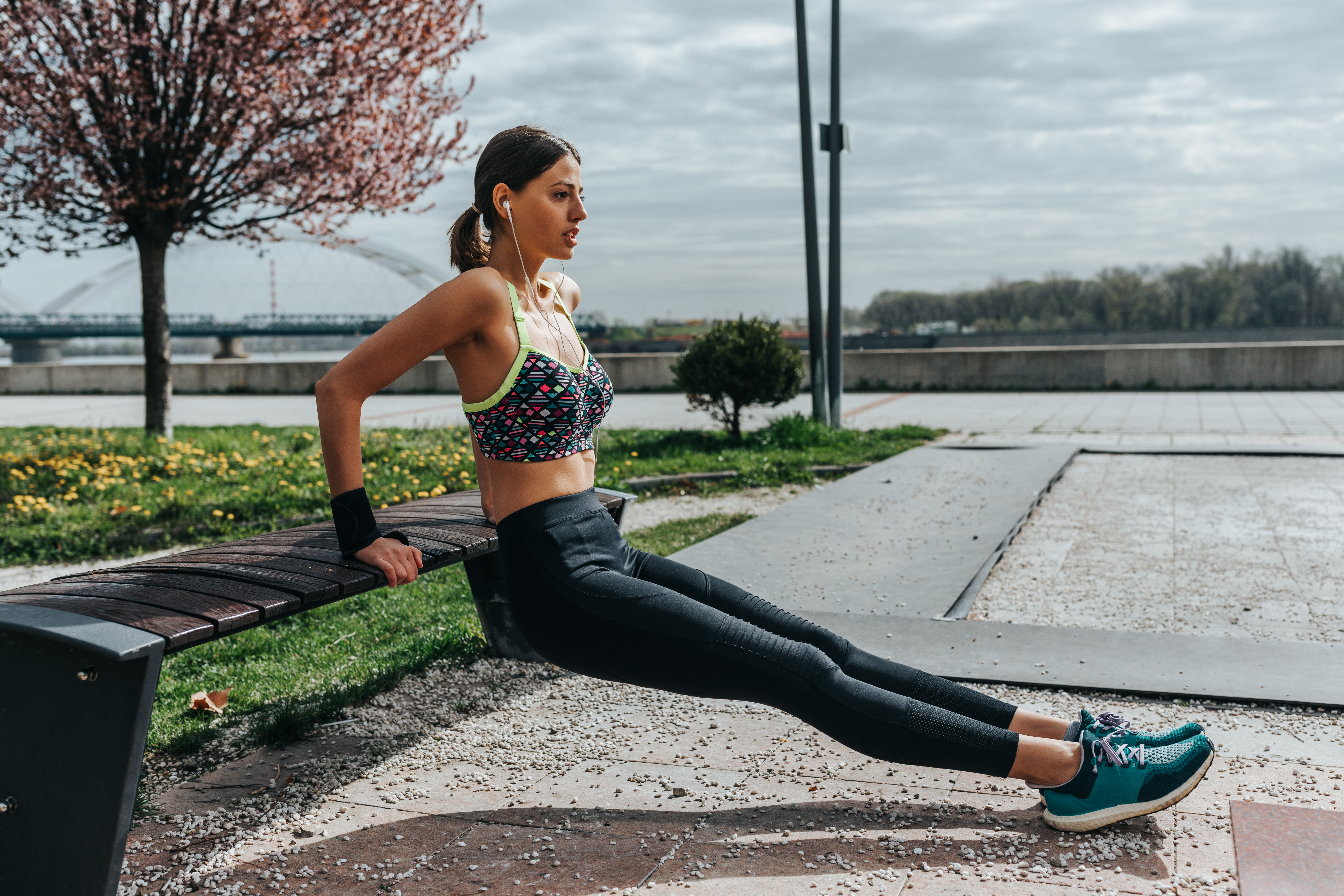 A young woman with brown hair in a ponytail, wearing a colorful sports bra and black leggings, exercises on a park bench.