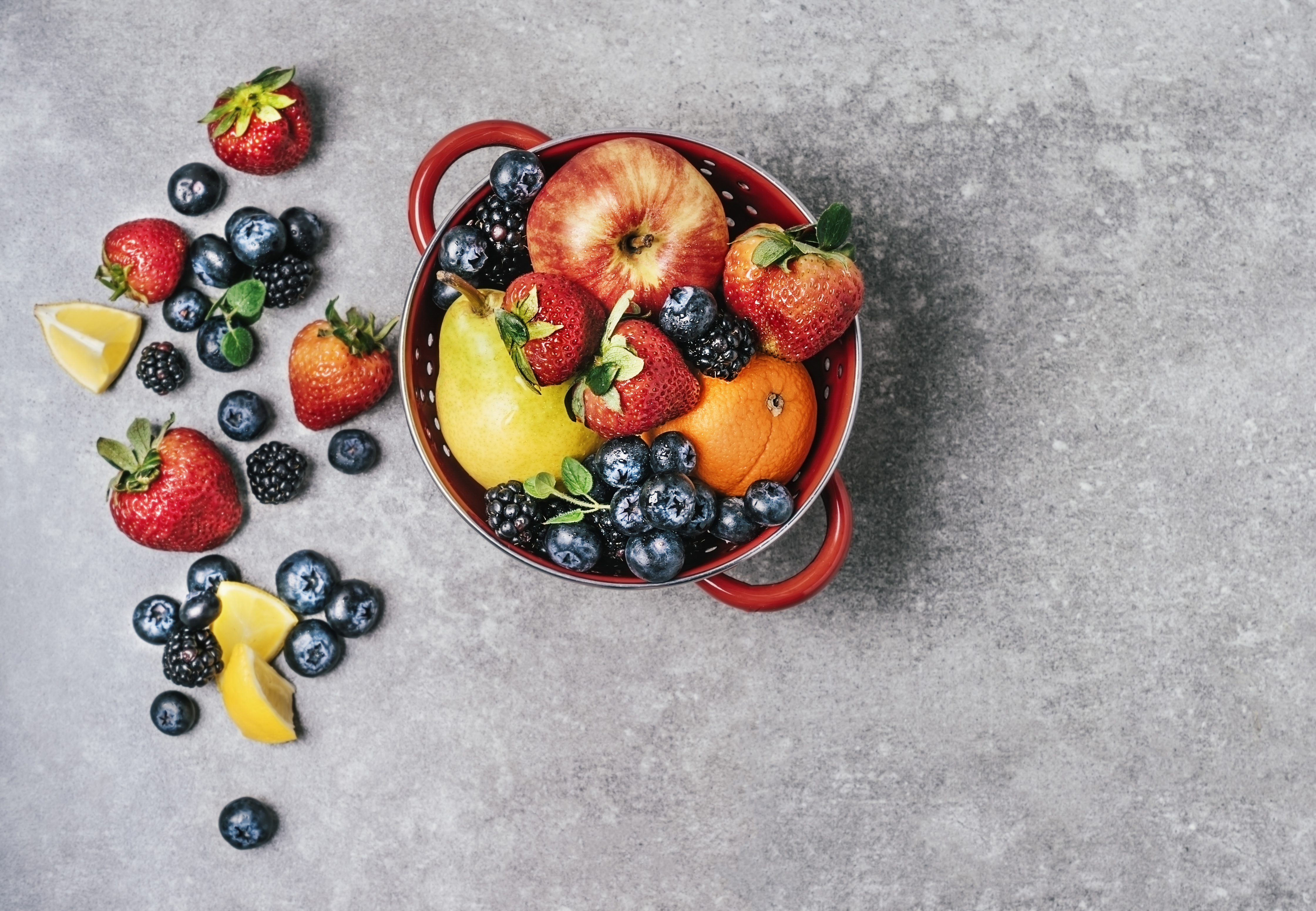 A colander overflowing with a variety of fresh fruits on a gray background.