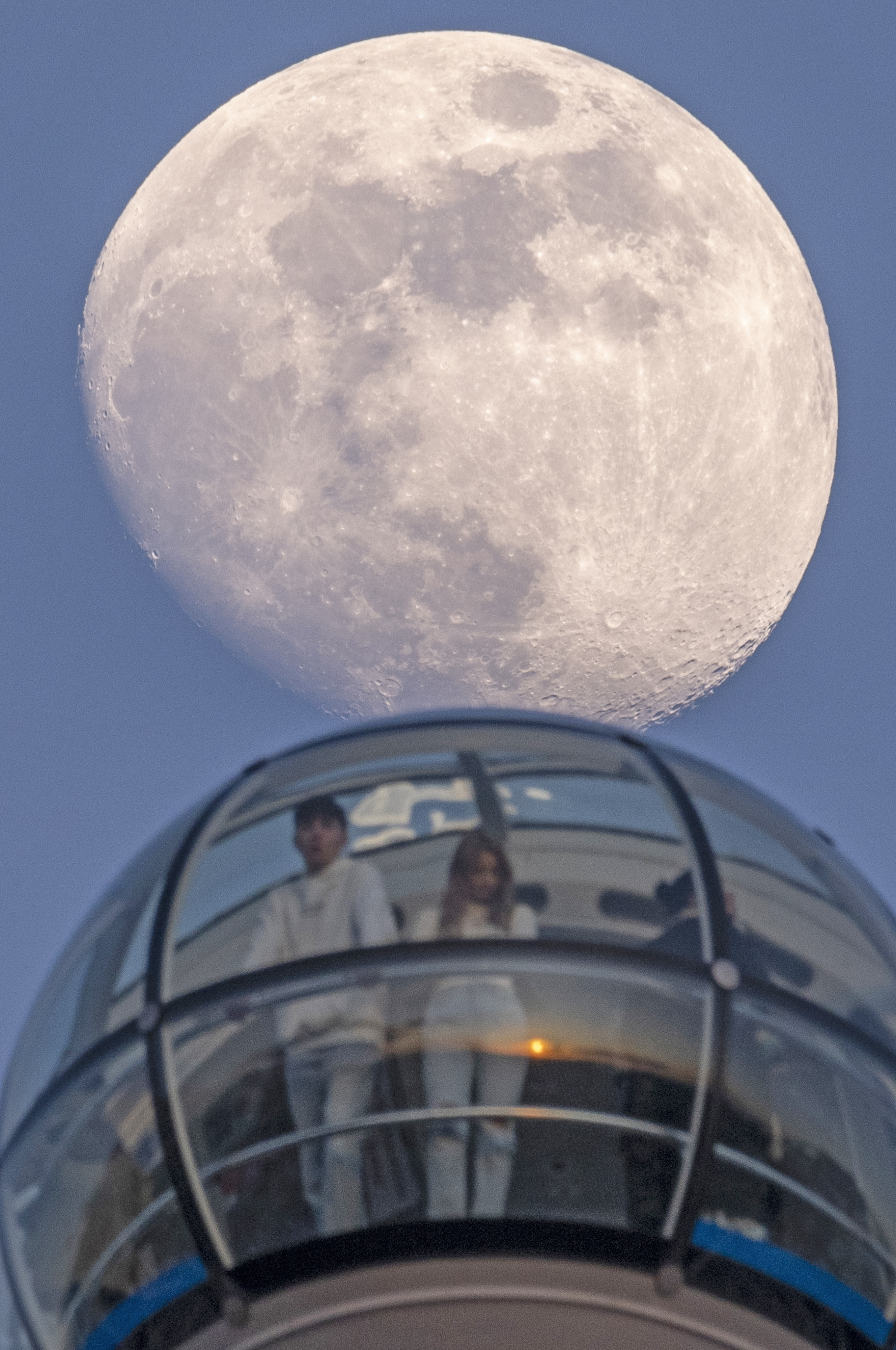 A large waxing moon is seen behind a pod of The London Eye, with people visible inside the pod.