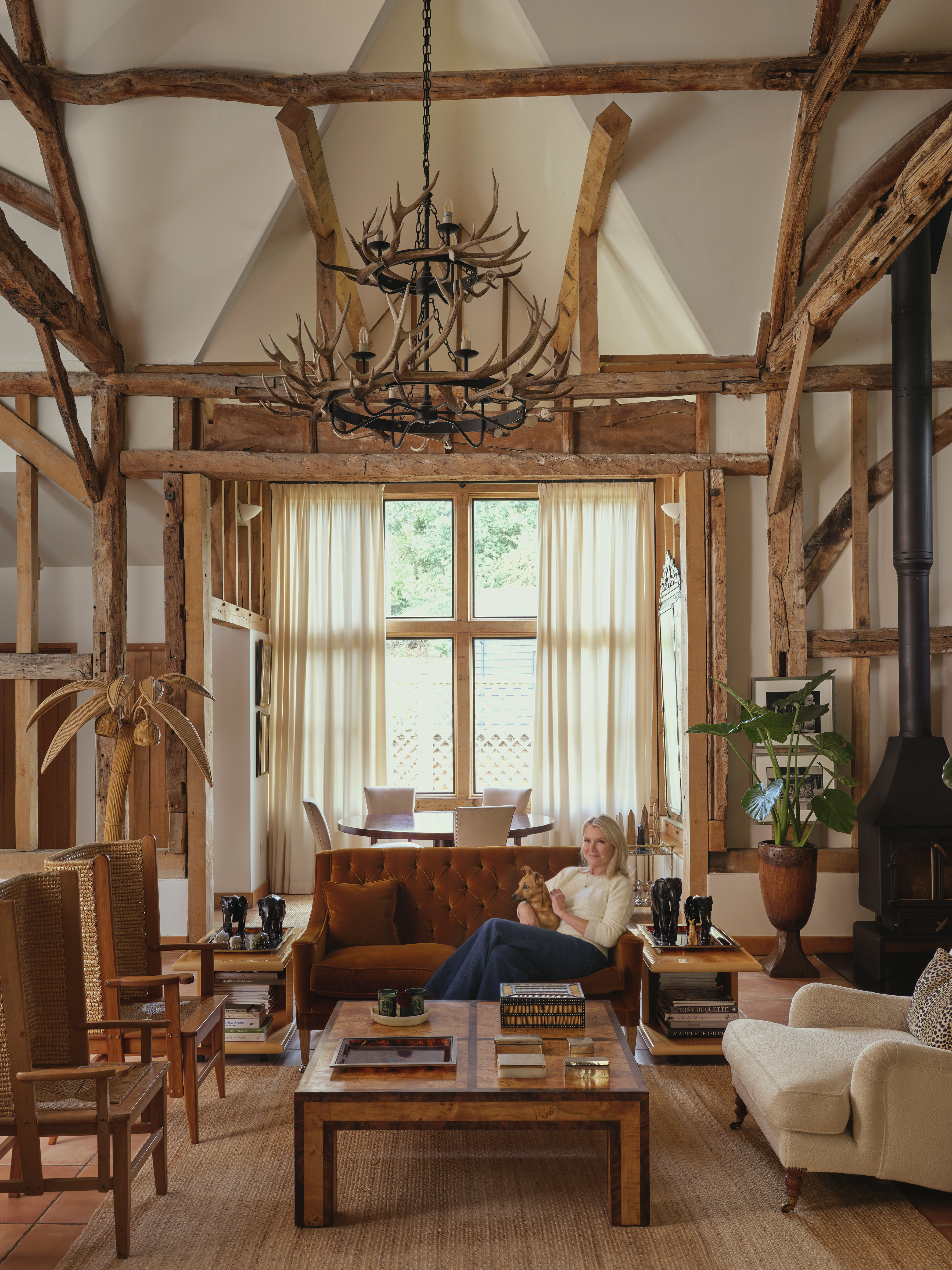 A woman sits on a brown sofa with a small dog, in a living room with exposed wooden beams and a large antler chandelier.
