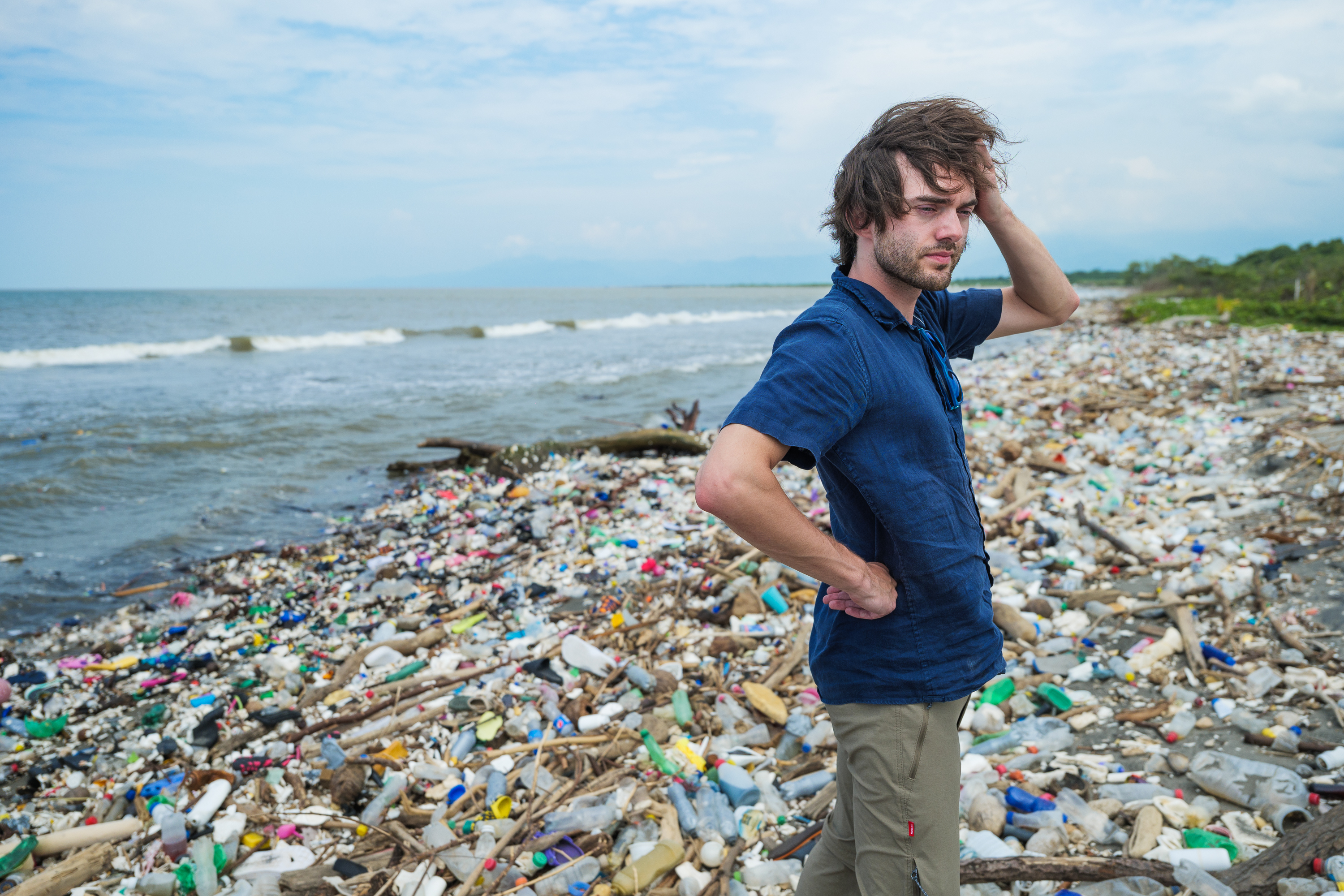 A man standing on a beach littered with plastic bottles and debris, looking distressed.