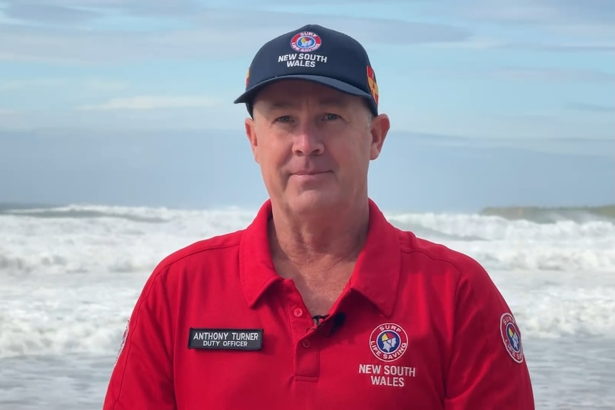 A man in red surf life saving uniform with a hat standing in front of large waves.