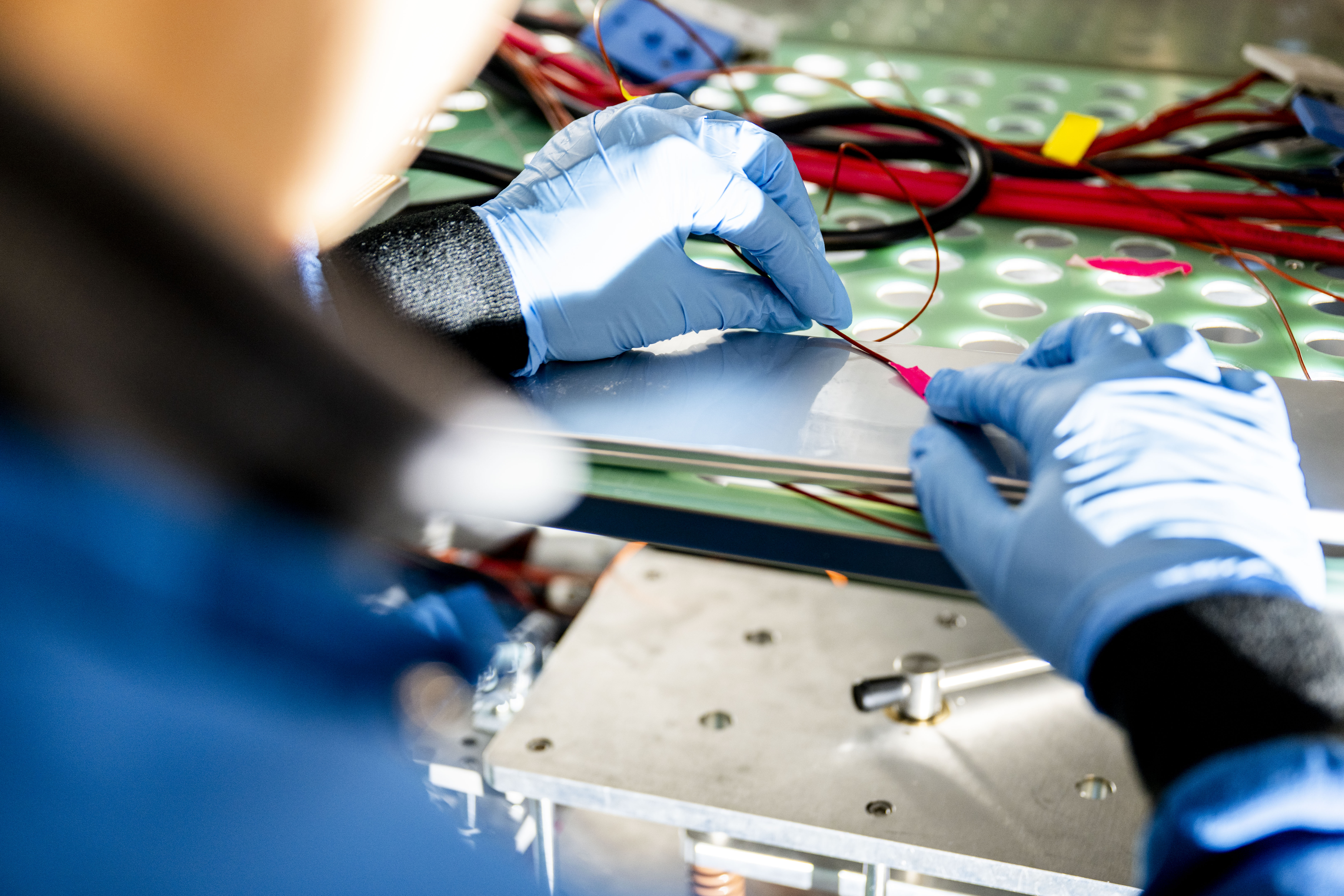 Gloved hands connect wires on a battery testing setup during electric vehicle research.