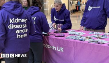 A group of eight women and one man standing in Queensgate Shopping Centre beside a sign that says Let's walk all over kidney disease. Six of the women and the man are wearing purple Kidney Research UK hoodies. The seventh woman who is in the middle of the group, is wearing a mayoral chain over her black jacket. The eighth woman, second on the left, is wearing a black coat over a grey top.
