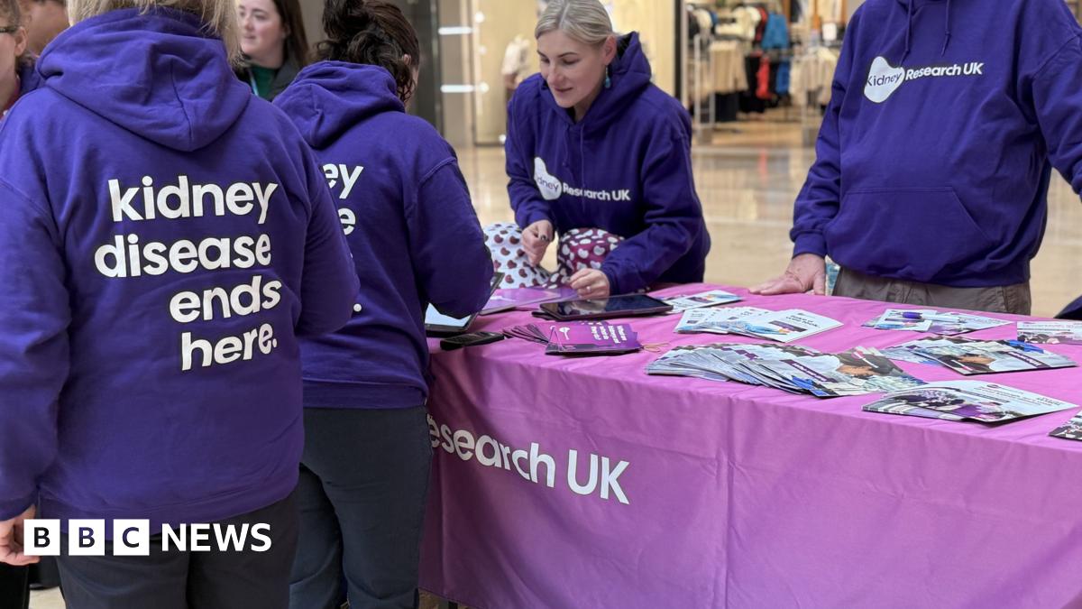 A group of eight women and one man standing in Queensgate Shopping Centre beside a sign that says Let's walk all over kidney disease. Six of the women and the man are wearing purple Kidney Research UK hoodies. The seventh woman who is in the middle of the group, is wearing a mayoral chain over her black jacket. The eighth woman, second on the left, is wearing a black coat over a grey top.
