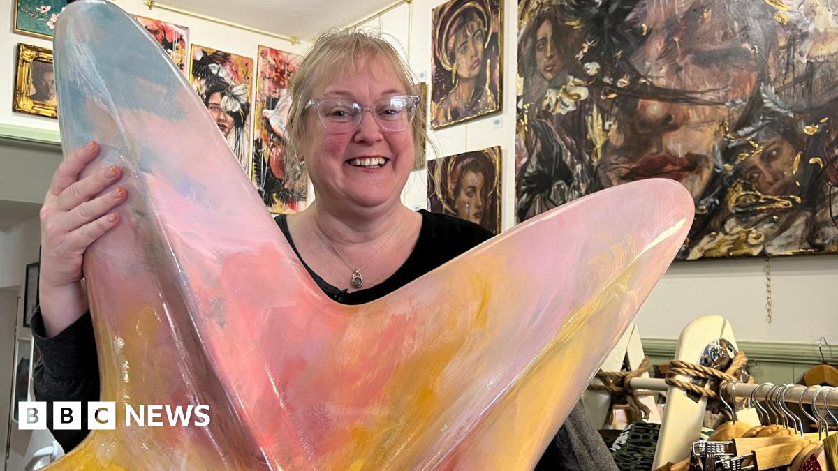 A woman smiles as she stands holding a large painted starfish sculpture in a gallery full of paintings. She has blonde hair pulled back and wears glasses and a black top. The starfish is painted in pinks, peaches and blues.
