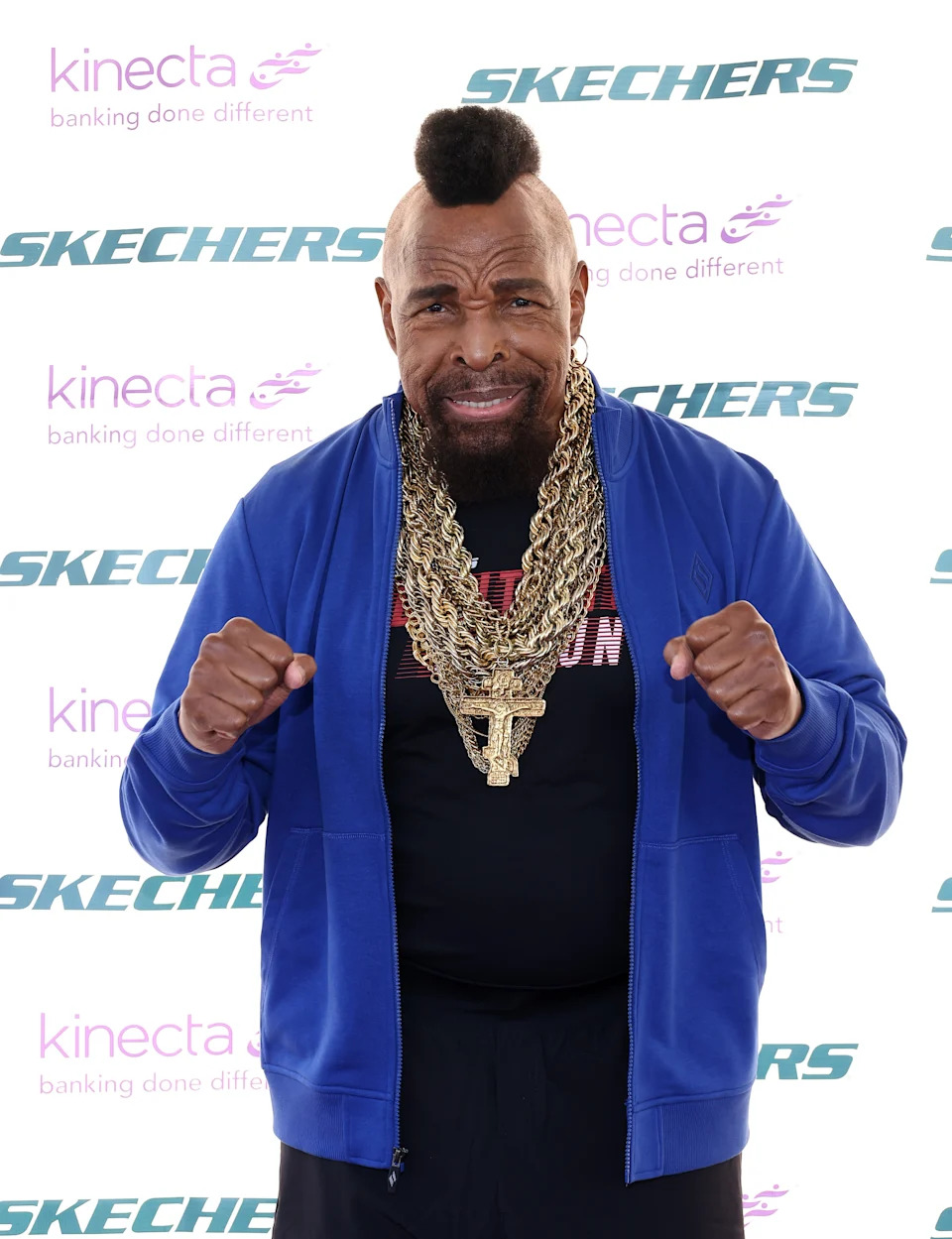 Mr. T in a blue jacket and multiple thick gold chains with a mohawk hairstyle, posing with fists up at a Skechers event backdrop