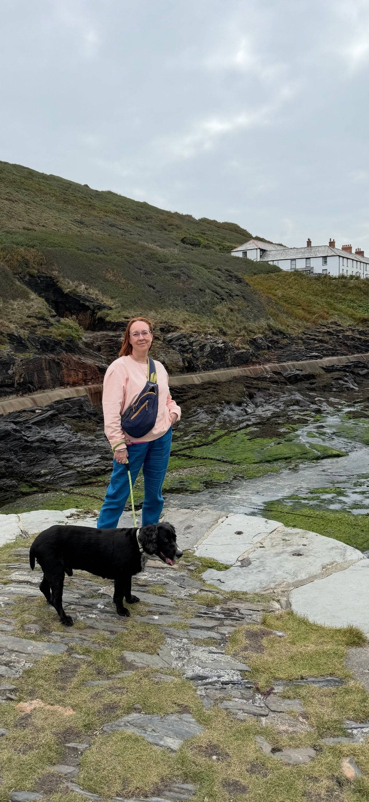 An individual is walking a black dog on a rocky trail near a small creek, with a house visible on a hillside in the background.