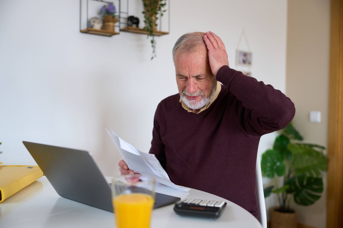 Worried senior man sitting at home, calculating domestic bills with a laptop and calculator, feeling stressed and frustrated over finances