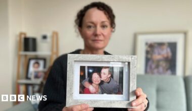 Emma Harley holds up a framed photograph of her and her brother Terry White.  Emma has dark curly hair, she is wearing a dark cardigan, and she is sitting on a green sofa surrounded by other photographs. In the photograph she is holding, she is wearing a red top and grimacing at the camera with her eyes closed. Her brother Terry has an arm around her shoulder and is giving her a kiss on the cheek. He has short shaved hair and is wearing a grey top.