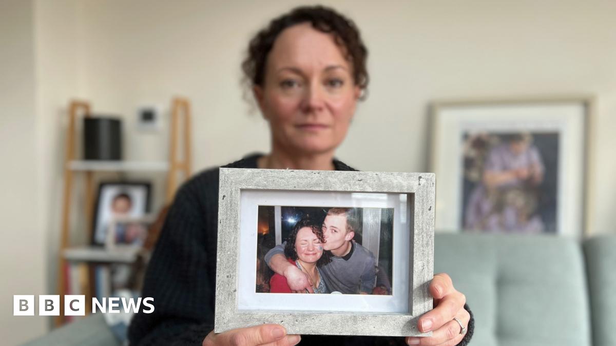 Emma Harley holds up a framed photograph of her and her brother Terry White.  Emma has dark curly hair, she is wearing a dark cardigan, and she is sitting on a green sofa surrounded by other photographs. In the photograph she is holding, she is wearing a red top and grimacing at the camera with her eyes closed. Her brother Terry has an arm around her shoulder and is giving her a kiss on the cheek. He has short shaved hair and is wearing a grey top.