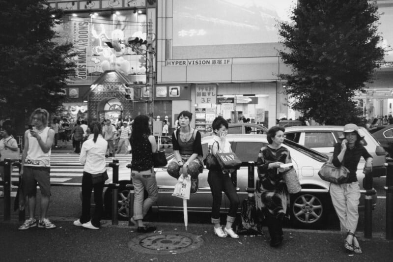 A group of people stand and sit on railings beside parked cars on a busy street in front of a large store with Japanese signs and cartoon decorations. Pedestrians and trees are visible in the background.