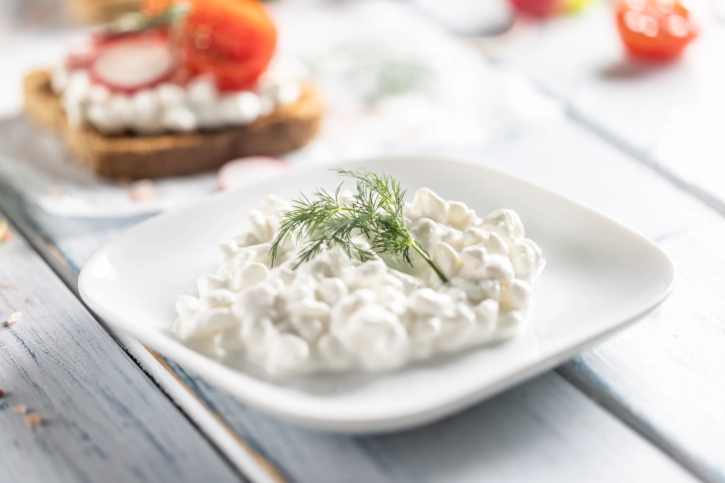 Cottage cheese with dill in a white bowl, with toasted bread topped with radish and cherry tomatoes in the background.