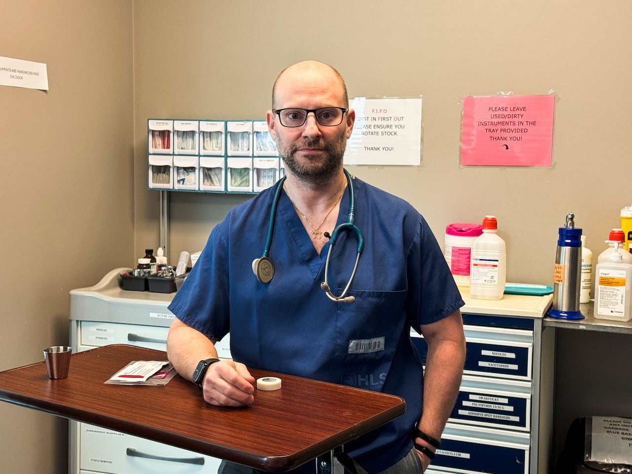 A man in scrubs with a stethoscope around his neck. He's standing in front of furniture storing medical equipment.