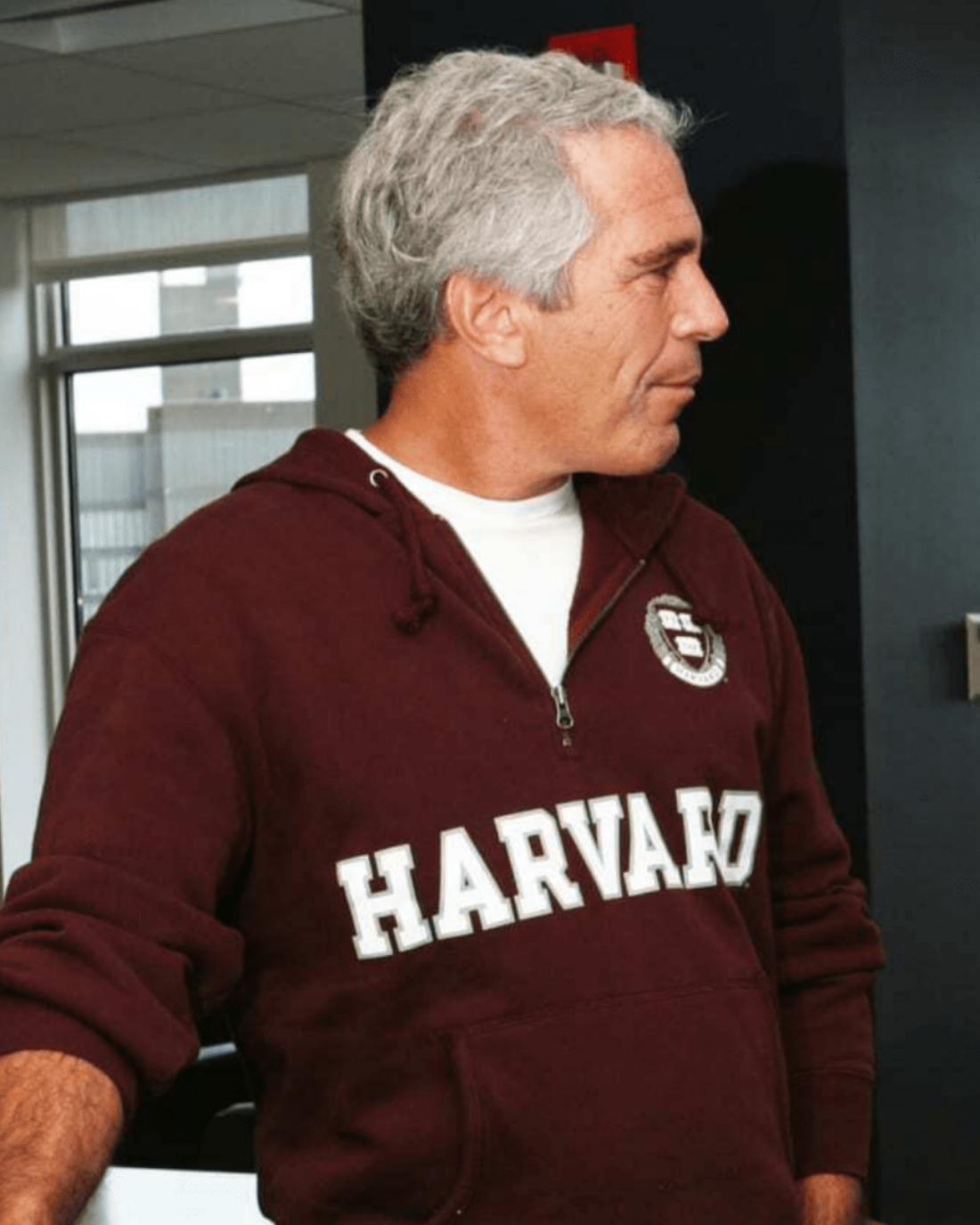 In this undated photo, Jeffrey Epstein is wearing a brick-red pullover hoodie that says "HARVARD" in white capital letters across the front. His head is turned toward the right side of the frame.
