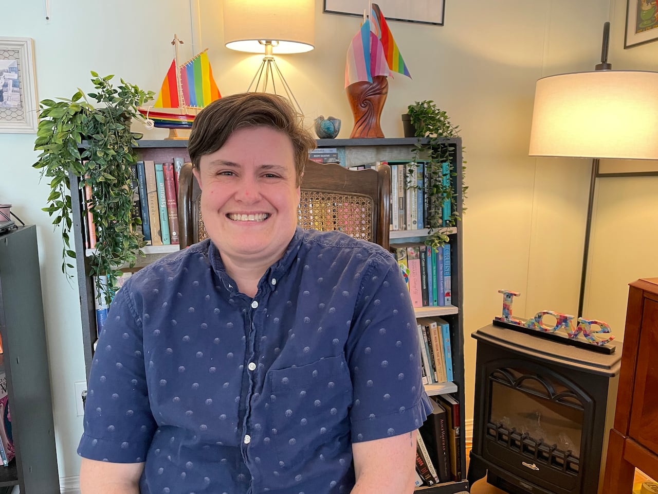 A non-binary person with short brown hair, wearing a blue button up shirt. They are smiling and sitting in their living room with a bookshelf behind them.