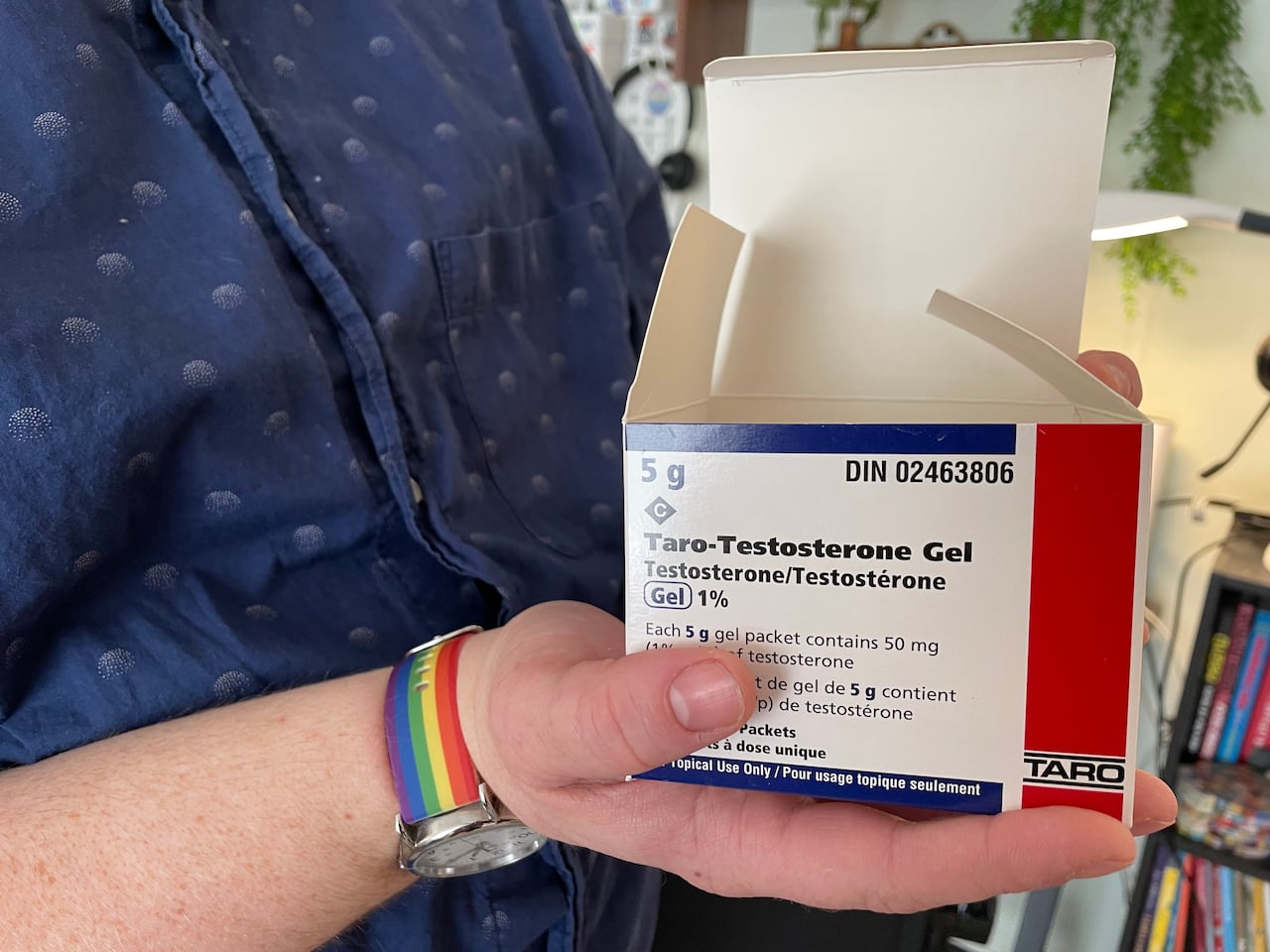 A hand holds out a box labelled "Taro-Testosterone Gel." You can also see the person is wearing a watch with a rainbow wristband and a blue button up shirt.
