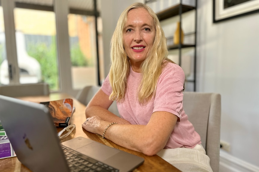 Woman with long blonde hair and pink t-shirt smiling at desk with computer.
