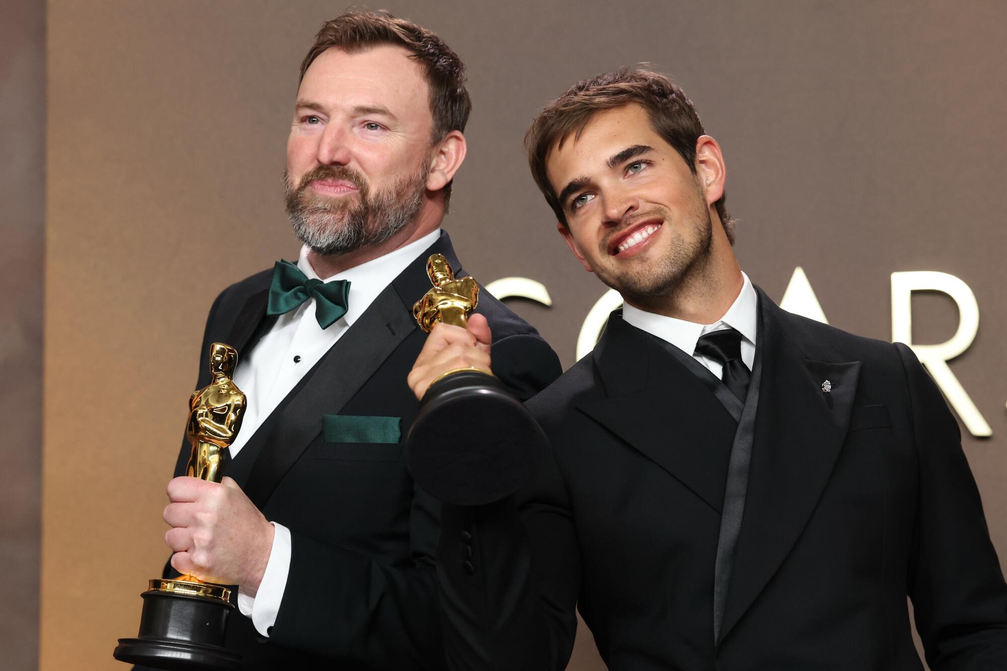 Two men in black tuxedos holding golden statuettes.