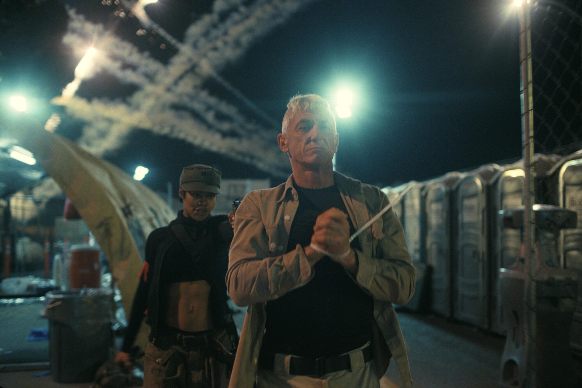 A man in military fatigues walking with zip ties around his hands as a woman in black walks behind him pointing a gun.