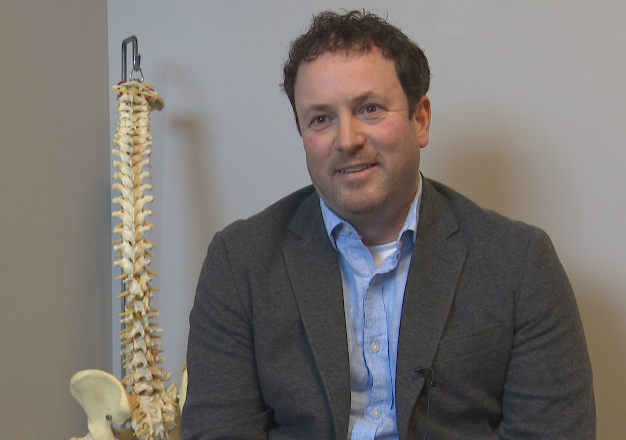 A man in a tweed jacket, wearing a blue shirt, sits in a chair next to a model of a spinal column, in a medical office.
