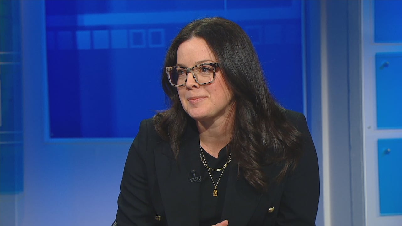 A woman in glasses and black clothing sits in front of a blue wall.
