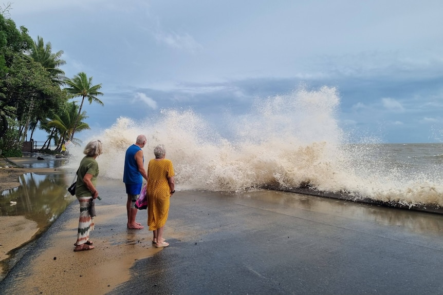 People stand on a beachside path as a wave splashes in front of them.