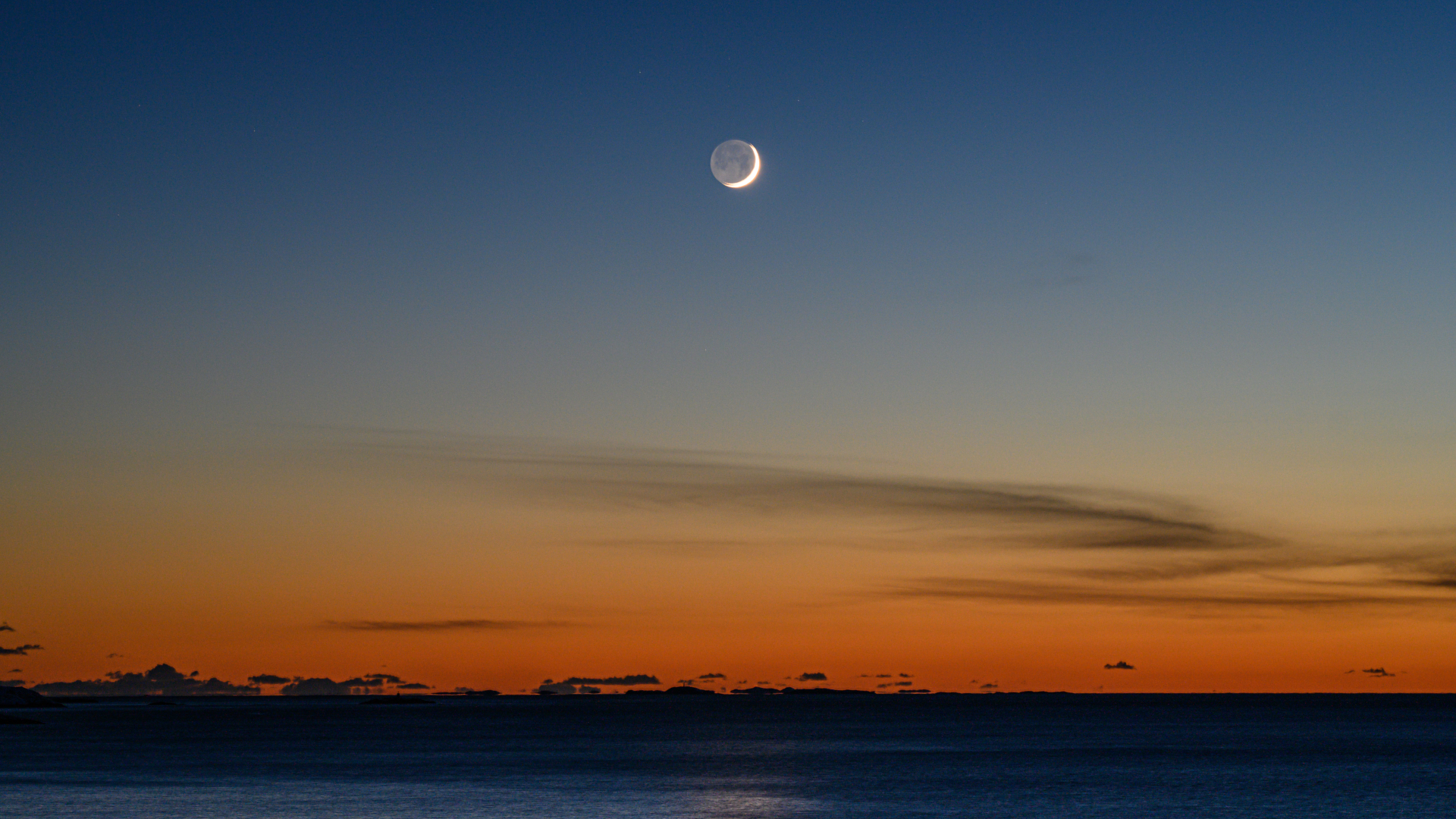 A photo of the thin crescent moon shining in the evening sky with its shadowed side lit by soft light reflected from Earth's surface. The orange glow of the setting sun is visible close to the horizon.