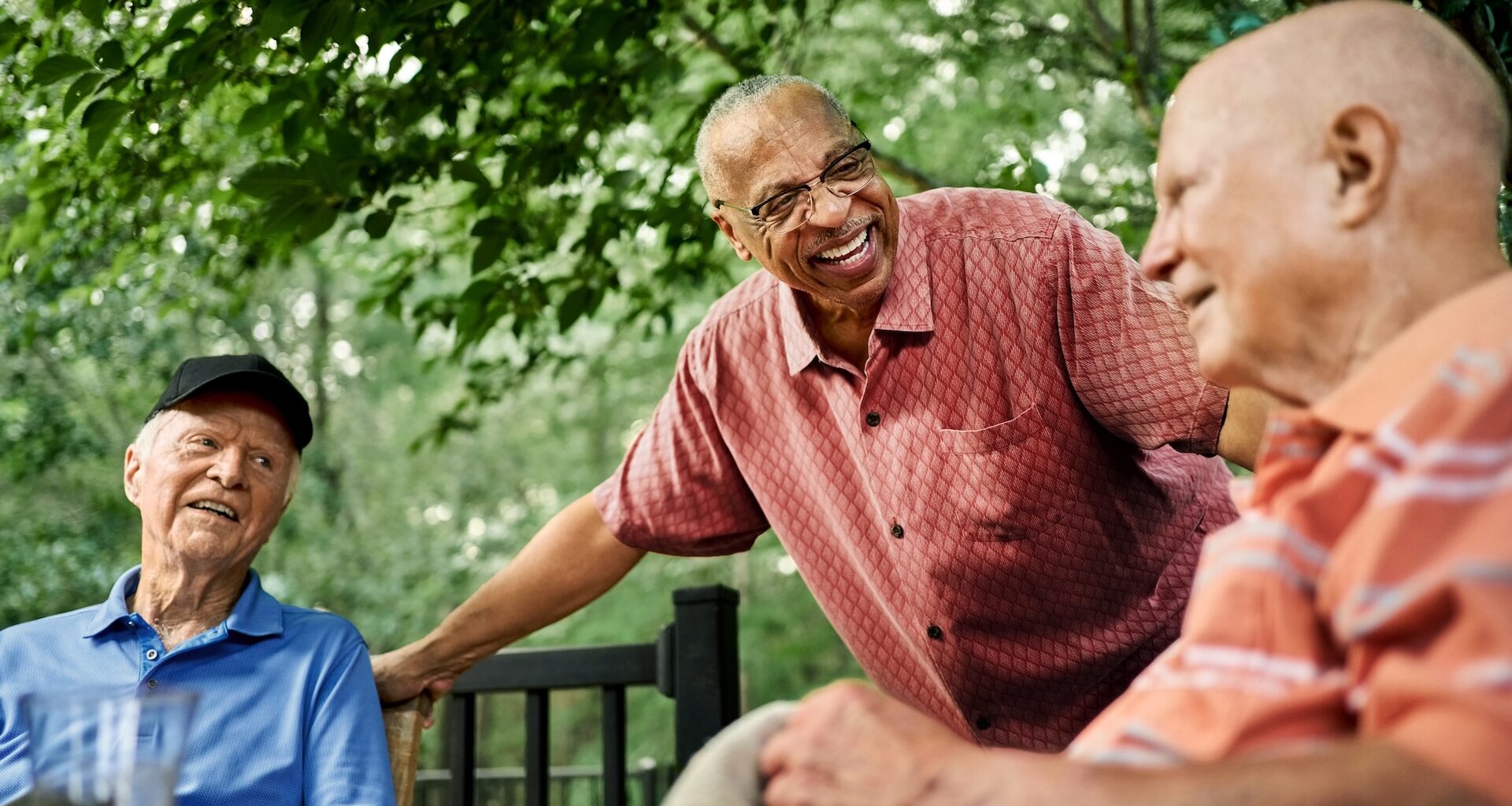 Group of happy senior male residents gathered around a table in the backyard of a retirement home, engaging in conversation and enjoying the serene outdoor space.