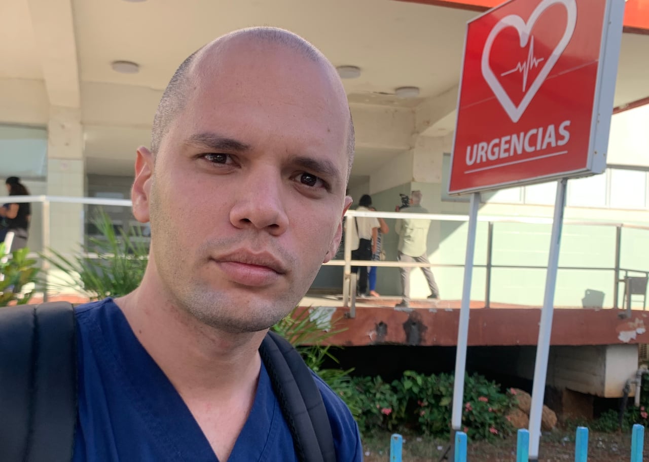 A man in a blue shirt looks straight ahead, in front of a hospital 