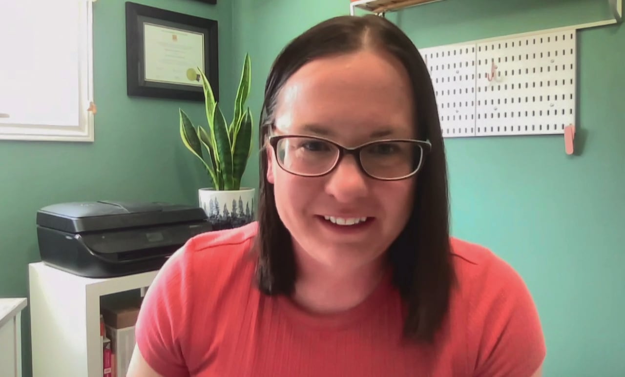 A brown-haired woman, wearing glasses, sits in a green medical office with a large, green aspidistra plant behind her