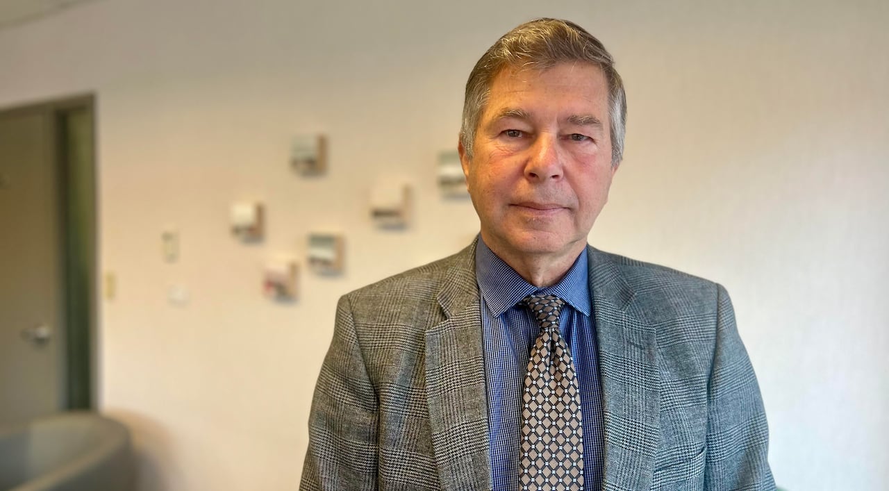 A man with grey hair, and a suit and tie, looks at the camera with a serious face, standing inside an office.  