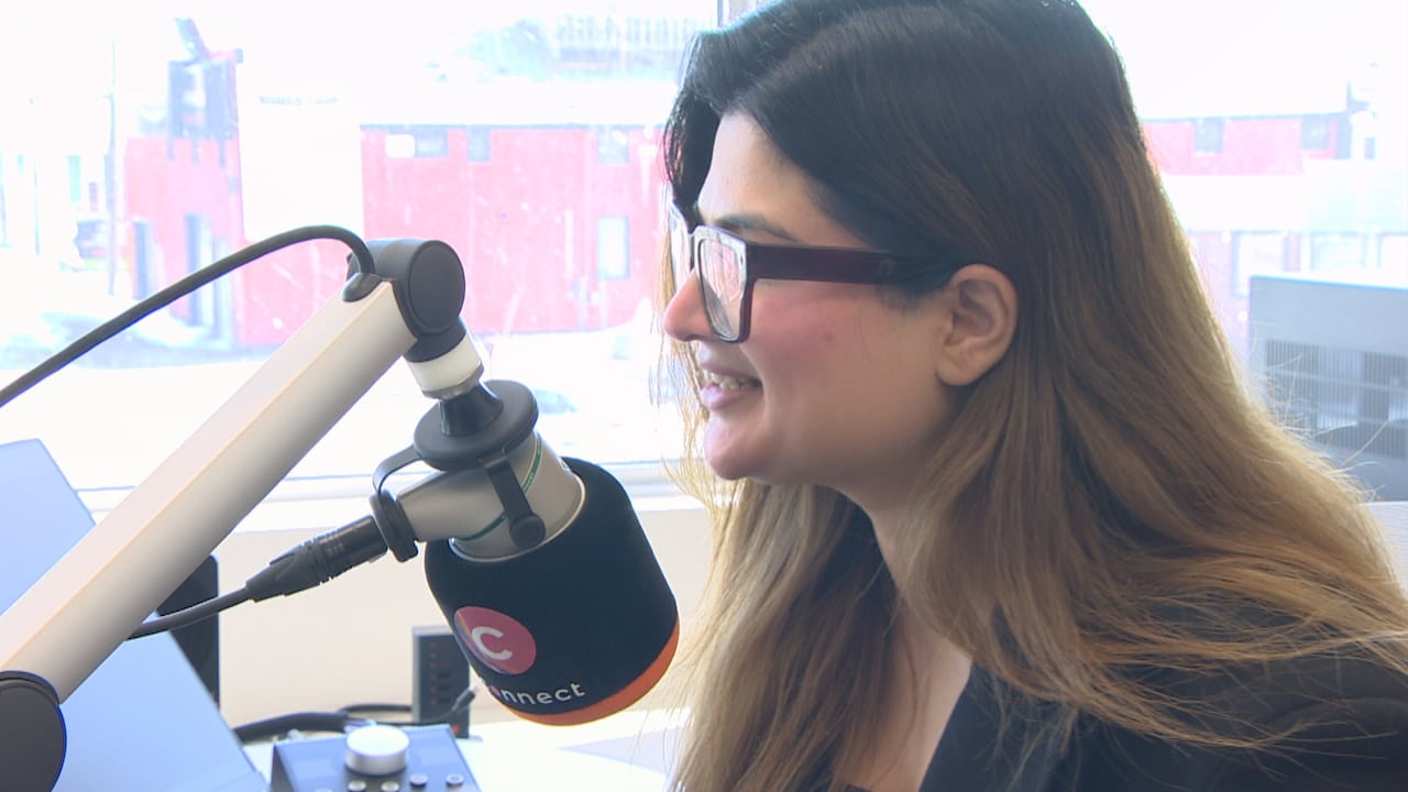 Woman with long dark hair and dark, oversized eyeglasses speaks into a black and orange microphone in a radio studio